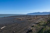 the sandy beach has a fence and dirt road near the water with a mountain in the distance