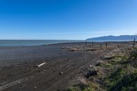 the sandy beach has a fence and dirt road near the water with a mountain in the distance