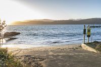 the sun setting behind the ocean on a beach with mountains in the background and a sign on a concrete pole at the end