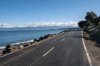 a lone empty asphalt road by the water and some rocks under a blue sky and clouds