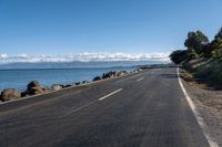 a lone empty asphalt road by the water and some rocks under a blue sky and clouds