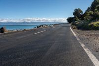 a lone empty asphalt road by the water and some rocks under a blue sky and clouds