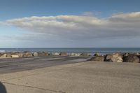 the skateboarder is carrying his board on the sidewalk next to the water and rocks