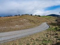 a dirt road curves over a hill with a blue sky above it and a hillside to the far