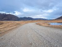 a gravel road running alongside an empty hill in the background is mountains with a lake in it