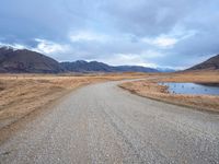 a gravel road running alongside an empty hill in the background is mountains with a lake in it