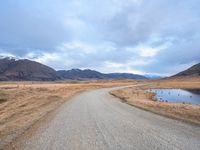 a gravel road running alongside an empty hill in the background is mountains with a lake in it