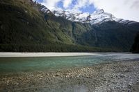 a body of water near a rocky hill next to a forest covered mountain range and snow capped peaks