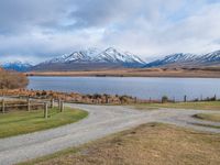 a park bench near a dirt road in an empty field near a lake and mountains