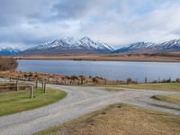 a park bench near a dirt road in an empty field near a lake and mountains