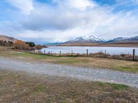 dirt path near a grassy field next to a pond and mountains with snow capped peaks