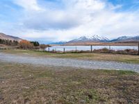 dirt path near a grassy field next to a pond and mountains with snow capped peaks