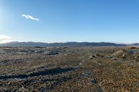 a field with a few rocks and a body of water in the distance, a mountains and blue sky