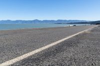 a paved street on the beach with water and mountains in the distance beyond it is a blue sky, and a white car parked nearby to the shore