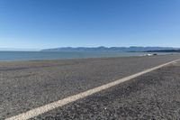 a paved street on the beach with water and mountains in the distance beyond it is a blue sky, and a white car parked nearby to the shore