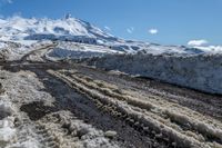 a road covered in mud next to snowy mountains in the distance with snow drifts and blue sky