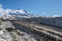 a road covered in mud next to snowy mountains in the distance with snow drifts and blue sky