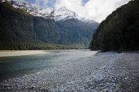 large stones with trees at the shore and snowy mountains in the background area of this photo