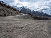 a dirt road going up a mountain side with snow covered mountains in the distance on both sides