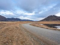 a dirt road passing through dry grass on the side of the mountain range with mountains in the distance