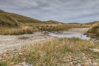 a wet road runs past some mountains and an empty stream near the grass, dirt and rocks