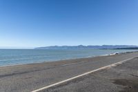 the side of an empty highway that is near water and mountains with blue skies on the background