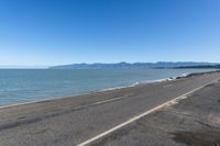 the side of an empty highway that is near water and mountains with blue skies on the background