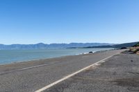 the side of an empty highway that is near water and mountains with blue skies on the background