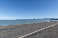 the side of an empty highway that is near water and mountains with blue skies on the background