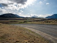 a dirt road leading to mountains with a sky in the background with white clouds above