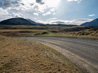 a dirt road leading to mountains with a sky in the background with white clouds above