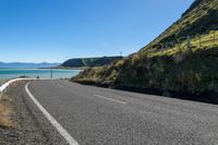 an empty road by the ocean near a hilly hill with many mountains in the background