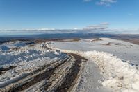 a view down to snow - covered road on a hilltop in the distance, and a man stands on top of it looking over the snow