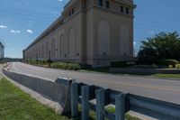 a street and two rows of cement barriers in front of a beige building, and an empty street with cars going by