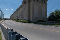 a street and two rows of cement barriers in front of a beige building, and an empty street with cars going by