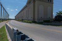 a street and two rows of cement barriers in front of a beige building, and an empty street with cars going by