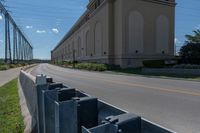 a street and two rows of cement barriers in front of a beige building, and an empty street with cars going by