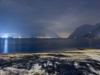 a bench sitting near the ocean during night with lights on mountains in the background on a cloudy day