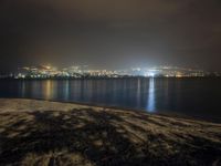 a bench sitting near the ocean during night with lights on mountains in the background on a cloudy day