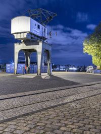 a tower on the side of a street with a train track running below it and some trees on a cobblestone road