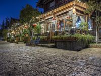 a cafe at night is lit up by lanterns and plants along the sidewalk or street