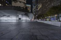 a city with a building in the background with some steps lit up at night as people sit on benches and tables near it