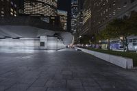 a city with a building in the background with some steps lit up at night as people sit on benches and tables near it