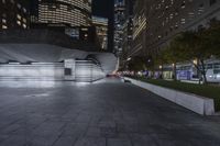 a city with a building in the background with some steps lit up at night as people sit on benches and tables near it