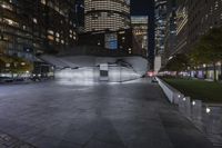 a city with a building in the background with some steps lit up at night as people sit on benches and tables near it