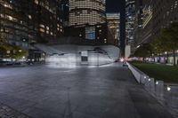 a city with a building in the background with some steps lit up at night as people sit on benches and tables near it