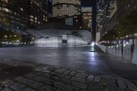 a city with a building in the background with some steps lit up at night as people sit on benches and tables near it