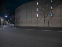 the empty street next to the building is dark at night with lit windows and trees in front of it