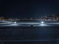 an empty parking lot near some street lights and buildings with lights reflecting on it and a river running under the roof