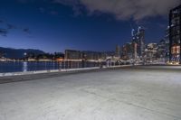 a paved area with water and buildings in the background at night as the sun sets